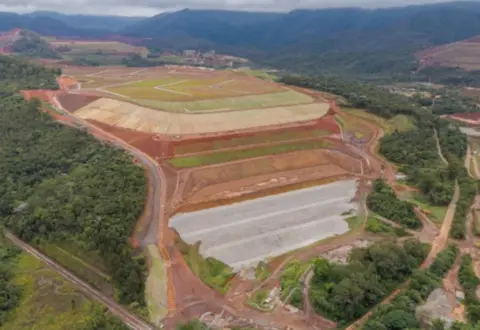 Barragem da Vale em Mariana tem classificação alterada após problemas em taludes e drenagem Segundo informações da ANM, a alteração ocorreu após serem identificadas mudanças no estado de conservação dos taludes, necessidade de revegetação e assoreamento em alguns dispositivos de drenagem superficial. Apesar da elevação da categoria, a agência informou que as ocorrências identificadas não estão associadas a um potencial comprometimento da segurança da estrutura. A Vale também afirma que Campo Grande permanece estável, segura e sem nível de alerta ou emergência. A Categoria de Risco é um indicador regulatório utilizado na fiscalização das barragens. O cálculo considera características técnicas, estado de conservação e o Plano de Segurança da Barragem. Pelos critérios da ANM, a categoria média corresponde, em regra, a pontuação superior a 40 e inferior a 80. Por isso, categoria de risco média não deve ser confundida com nível de emergência. A mudança registrada para Campo Grande não significa, por si só, que a barragem esteja em situação de emergência. Vale já iniciou medidas corretivas A ANM determinou que a Vale adotasse medidas para corrigir as condições encontradas. Em uma inspeção regular reportada pela própria mineradora à agência em 17 de julho, a empresa informou que as intervenções já estavam sendo executadas. A Vale declarou que Campo Grande continua sendo monitorada durante 24 horas por dia, sete dias por semana, pelos Centros de Monitoramento Geotécnico da companhia. A mineradora sustenta ainda que, durante o período posterior à descaracterização, a Categoria de Risco pode apresentar variações relacionadas a necessidades rotineiras de manutenção e reforça que essa classificação não equivale a uma avaliação direta da estabilidade. O próprio histórico de Campo Grande após as obras registra intervenções desse tipo. Relatório da Vale informa que, já em janeiro de 2026, foram realizadas limpeza de dispositivos de drenagem, manutenção de acessos e adequações pontuais decorrentes das chuvas. Obras de descaracterização terminaram em dezembro A Campo Grande foi construída pelo método de alteamento a montante e teve as obras de descaracterização concluídas em dezembro de 2025. A Vale anunciou na ocasião que era a 19ª estrutura do tipo a ter as intervenções concluídas dentro de seu programa. Os trabalhos envolveram a retirada do alteamento a montante, reconfiguração da estrutura, construção de reforços, impermeabilização e adequações no sistema de drenagem. A descaracterização modifica a estrutura para que ela deixe de funcionar como uma barragem de rejeitos nos moldes anteriores. No caso de Campo Grande, porém, o processo não envolveu a retirada do rejeito armazenado, conforme informações divulgadas pela própria Vale. Em relatório posterior às obras, a empresa afirmou entender que Campo Grande não se enquadra mais no conceito legal de barragem por não operar como estrutura de contenção de sedimentos ou rejeitos. A Vale também informou ter protocolado junto à Fundação Estadual do Meio Ambiente (Feam) pedido para o descadastramento da estrutura. Monitoramento continua após a descaracterização A conclusão das obras não encerra imediatamente o acompanhamento da estrutura. Campo Grande permanece no período de monitoramento previsto após a descaracterização. Relatório da ANM sobre as barragens a montante registra dezembro de 2025 como início desse período e dezembro de 2027 como previsão de término do acompanhamento da Campo Grande. Ao anunciar a conclusão das obras, a própria Vale informou que a estrutura permaneceria monitorada por pelo menos dois anos, enquanto a documentação e as evidências técnicas da descaracterização seriam submetidas aos órgãos competentes. É justamente durante esse período posterior às intervenções que ocorreu a mudança da Categoria de Risco identificada pela ANM. Campo Grande tem 94 metros de altura A estrutura tem 94 metros de altura, dimensão próxima à de um edifício de aproximadamente 30 andares, e reservatório de 20,1 milhões de metros cúbicos. Apesar da Categoria de Risco média, Campo Grande é classificada com Dano Potencial Associado (DPA) alto. Os dois indicadores medem aspectos diferentes. O DPA considera as possíveis consequências de um eventual rompimento, incluindo perdas de vidas, impactos ambientais e efeitos socioeconômicos. Já a Categoria de Risco considera fatores ligados às características técnicas, conservação e gestão de segurança. De acordo com os dados apresentados no Sistema Nacional de Informações sobre Segurança de Barragens, a área potencialmente afetada a jusante possui ocupação permanente, com estimativa de que entre mil e 5 mil pessoas poderiam ser atingidas em um cenário hipotético de ruptura. Mudança ocorre dias após embargo de outra estrutura da Vale A alteração na classificação da Campo Grande ocorre em um período de outras decisões da ANM envolvendo estruturas da Vale na Região Central de Minas. Em 28 de agosto, a agência determinou o embargo da barragem Maravilhas II, na Mina do Pico, em Itabirito, impedindo novos depósitos de rejeitos até a regularização de documentos relacionados à segurança. Como mostrou o Mais Minas, o embargo da barragem Maravilhas II ocorreu após a ANM identificar que documentos apresentados pela Vale não refletiam as condições da estrutura depois de obras realizadas no local. Naquele caso, porém, a agência esclareceu que a medida não foi motivada pela identificação de uma nova anomalia física. Maravilhas II havia deixado o nível 1 de emergência em janeiro e estava inoperante por planejamento operacional quando ocorreu o embargo. Os dois casos, portanto, têm fundamentos diferentes: em Campo Grande, houve alteração da Categoria de Risco relacionada ao estado de conservação; em Maravilhas II, a medida decorreu de pendências na atualização da documentação exigida pela ANM. O sistema público da agência permite consultar a classificação atualizada das barragens, localização e histórico de informações de segurança.