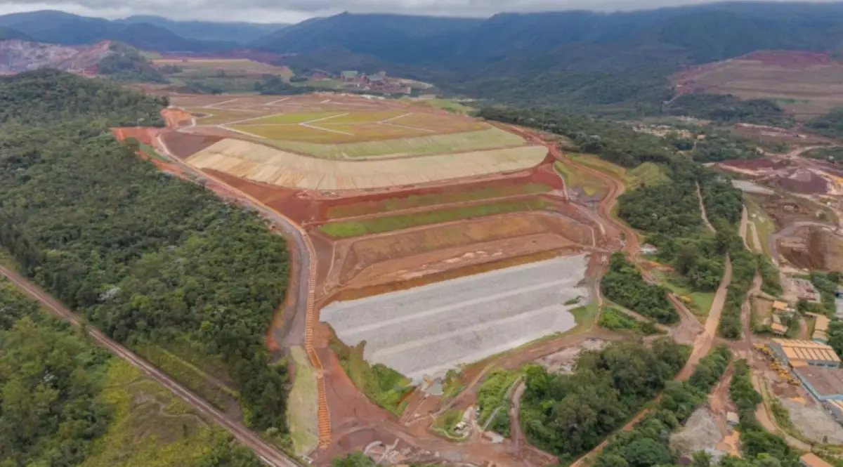 Barragem da Vale em Mariana tem classificação alterada após problemas em taludes e drenagem Segundo informações da ANM, a alteração ocorreu após serem identificadas mudanças no estado de conservação dos taludes, necessidade de revegetação e assoreamento em alguns dispositivos de drenagem superficial. Apesar da elevação da categoria, a agência informou que as ocorrências identificadas não estão associadas a um potencial comprometimento da segurança da estrutura. A Vale também afirma que Campo Grande permanece estável, segura e sem nível de alerta ou emergência. A Categoria de Risco é um indicador regulatório utilizado na fiscalização das barragens. O cálculo considera características técnicas, estado de conservação e o Plano de Segurança da Barragem. Pelos critérios da ANM, a categoria média corresponde, em regra, a pontuação superior a 40 e inferior a 80. Por isso, categoria de risco média não deve ser confundida com nível de emergência. A mudança registrada para Campo Grande não significa, por si só, que a barragem esteja em situação de emergência. Vale já iniciou medidas corretivas A ANM determinou que a Vale adotasse medidas para corrigir as condições encontradas. Em uma inspeção regular reportada pela própria mineradora à agência em 17 de julho, a empresa informou que as intervenções já estavam sendo executadas. A Vale declarou que Campo Grande continua sendo monitorada durante 24 horas por dia, sete dias por semana, pelos Centros de Monitoramento Geotécnico da companhia. A mineradora sustenta ainda que, durante o período posterior à descaracterização, a Categoria de Risco pode apresentar variações relacionadas a necessidades rotineiras de manutenção e reforça que essa classificação não equivale a uma avaliação direta da estabilidade. O próprio histórico de Campo Grande após as obras registra intervenções desse tipo. Relatório da Vale informa que, já em janeiro de 2026, foram realizadas limpeza de dispositivos de drenagem, manutenção de acessos e adequações pontuais decorrentes das chuvas. Obras de descaracterização terminaram em dezembro A Campo Grande foi construída pelo método de alteamento a montante e teve as obras de descaracterização concluídas em dezembro de 2025. A Vale anunciou na ocasião que era a 19ª estrutura do tipo a ter as intervenções concluídas dentro de seu programa. Os trabalhos envolveram a retirada do alteamento a montante, reconfiguração da estrutura, construção de reforços, impermeabilização e adequações no sistema de drenagem. A descaracterização modifica a estrutura para que ela deixe de funcionar como uma barragem de rejeitos nos moldes anteriores. No caso de Campo Grande, porém, o processo não envolveu a retirada do rejeito armazenado, conforme informações divulgadas pela própria Vale. Em relatório posterior às obras, a empresa afirmou entender que Campo Grande não se enquadra mais no conceito legal de barragem por não operar como estrutura de contenção de sedimentos ou rejeitos. A Vale também informou ter protocolado junto à Fundação Estadual do Meio Ambiente (Feam) pedido para o descadastramento da estrutura. Monitoramento continua após a descaracterização A conclusão das obras não encerra imediatamente o acompanhamento da estrutura. Campo Grande permanece no período de monitoramento previsto após a descaracterização. Relatório da ANM sobre as barragens a montante registra dezembro de 2025 como início desse período e dezembro de 2027 como previsão de término do acompanhamento da Campo Grande. Ao anunciar a conclusão das obras, a própria Vale informou que a estrutura permaneceria monitorada por pelo menos dois anos, enquanto a documentação e as evidências técnicas da descaracterização seriam submetidas aos órgãos competentes. É justamente durante esse período posterior às intervenções que ocorreu a mudança da Categoria de Risco identificada pela ANM. Campo Grande tem 94 metros de altura A estrutura tem 94 metros de altura, dimensão próxima à de um edifício de aproximadamente 30 andares, e reservatório de 20,1 milhões de metros cúbicos. Apesar da Categoria de Risco média, Campo Grande é classificada com Dano Potencial Associado (DPA) alto. Os dois indicadores medem aspectos diferentes. O DPA considera as possíveis consequências de um eventual rompimento, incluindo perdas de vidas, impactos ambientais e efeitos socioeconômicos. Já a Categoria de Risco considera fatores ligados às características técnicas, conservação e gestão de segurança. De acordo com os dados apresentados no Sistema Nacional de Informações sobre Segurança de Barragens, a área potencialmente afetada a jusante possui ocupação permanente, com estimativa de que entre mil e 5 mil pessoas poderiam ser atingidas em um cenário hipotético de ruptura. Mudança ocorre dias após embargo de outra estrutura da Vale A alteração na classificação da Campo Grande ocorre em um período de outras decisões da ANM envolvendo estruturas da Vale na Região Central de Minas. Em 28 de agosto, a agência determinou o embargo da barragem Maravilhas II, na Mina do Pico, em Itabirito, impedindo novos depósitos de rejeitos até a regularização de documentos relacionados à segurança. Como mostrou o Mais Minas, o embargo da barragem Maravilhas II ocorreu após a ANM identificar que documentos apresentados pela Vale não refletiam as condições da estrutura depois de obras realizadas no local. Naquele caso, porém, a agência esclareceu que a medida não foi motivada pela identificação de uma nova anomalia física. Maravilhas II havia deixado o nível 1 de emergência em janeiro e estava inoperante por planejamento operacional quando ocorreu o embargo. Os dois casos, portanto, têm fundamentos diferentes: em Campo Grande, houve alteração da Categoria de Risco relacionada ao estado de conservação; em Maravilhas II, a medida decorreu de pendências na atualização da documentação exigida pela ANM. O sistema público da agência permite consultar a classificação atualizada das barragens, localização e histórico de informações de segurança.
